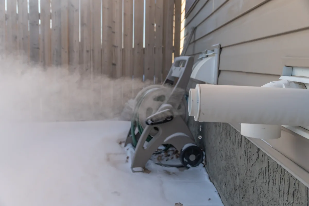 Snow drifted against the white plastic intake and exhaust vents on the side wall of a house