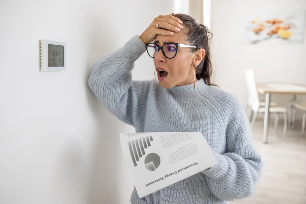 Homeowner shocked by a high winter energy bill while standing beside the thermostat